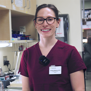 Smiling woman in maroon scrubs and glasses, wearing a name badge and microphone, stands in a clinic or medical office with shelves, supplies, and a paper towel dispenser visible in the background.