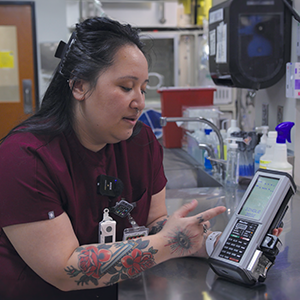A person in medical scrubs uses a syringe pump at a stainless steel workstation in a clinical or laboratory setting.