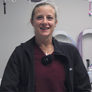 A person wearing a maroon shirt and black jacket is smiling indoors, standing in a room with medical or veterinary equipment visible in the background.