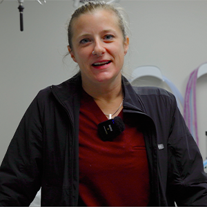 A woman with blonde hair tied back, wearing a maroon top and black jacket, stands indoors with medical equipment in the background. She has a small microphone clipped to her jacket.