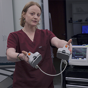 A medical professional in burgundy scrubs holds defibrillator paddles in a clinical setting, with a heart monitor displaying vital signs in the background.
