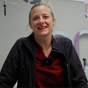 A smiling person in scrubs and a black jacket stands in a medical or veterinary clinic, with medical equipment visible in the background.