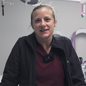 A person with light hair pulled back, wearing a dark jacket and a maroon shirt, stands in a medical or clinical setting with equipment visible in the background.