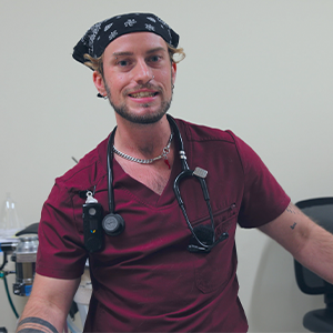 A smiling person in maroon scrubs and a black bandana, wearing a stethoscope around their neck, stands in a medical office setting.