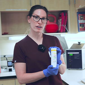 A woman in medical scrubs and gloves holds a urine test strip container in a lab setting, with equipment and supplies visible in the background.