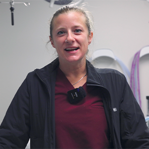 A woman wearing a maroon shirt and black jacket smiles in a medical setting, with medical equipment and a stethoscope visible in the background. She has a microphone clipped to her jacket.