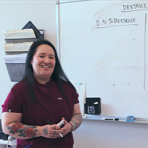 A smiling healthcare professional in a maroon scrub top stands in front of a whiteboard that reads DEXTROSE and 2.5% DEXTROSE.