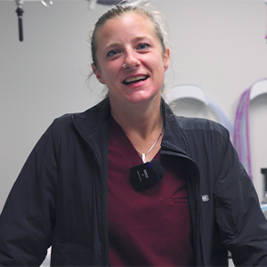 A woman in scrubs and a dark jacket stands indoors, smiling at the camera. She wears a microphone on her jacket, and medical equipment is visible in the background.