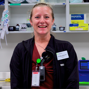 A smiling woman in a maroon scrub top and black jacket, with a hospital ID badge and equipment clipped to her chest, standing in a medical storage room.